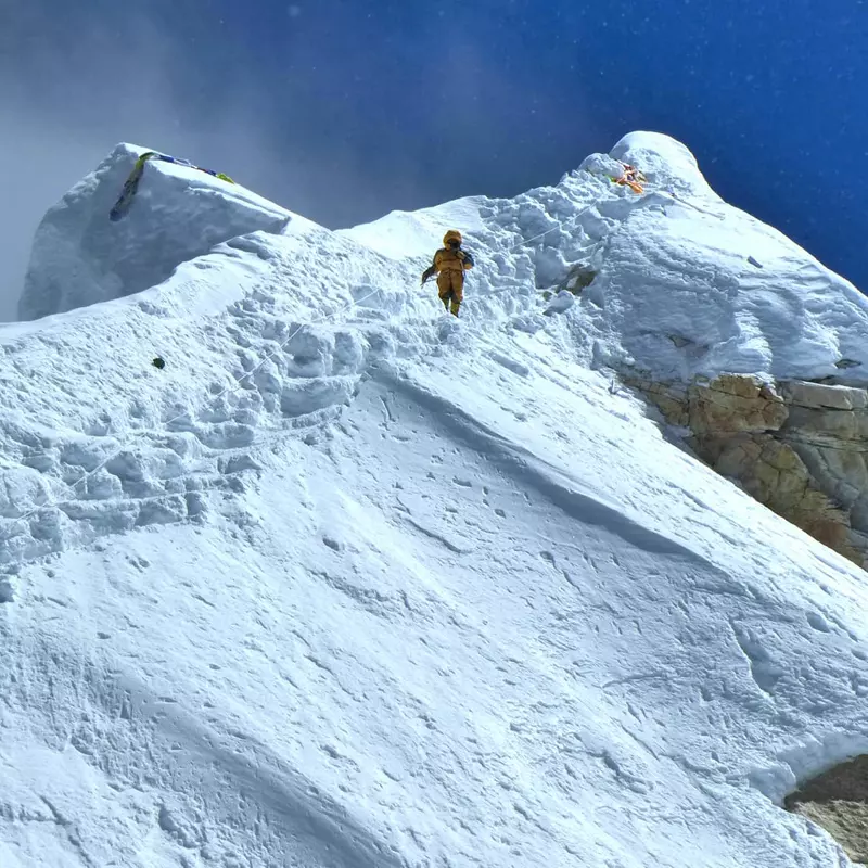 Een bergbeklimmer in een geel pak beklimt een steile, met sneeuw bedekte bergkam onder een strakblauwe hemel, terwijl hij diepe voetafdrukken achterlaat en op de top twee kleine tenten te zien zijn.
